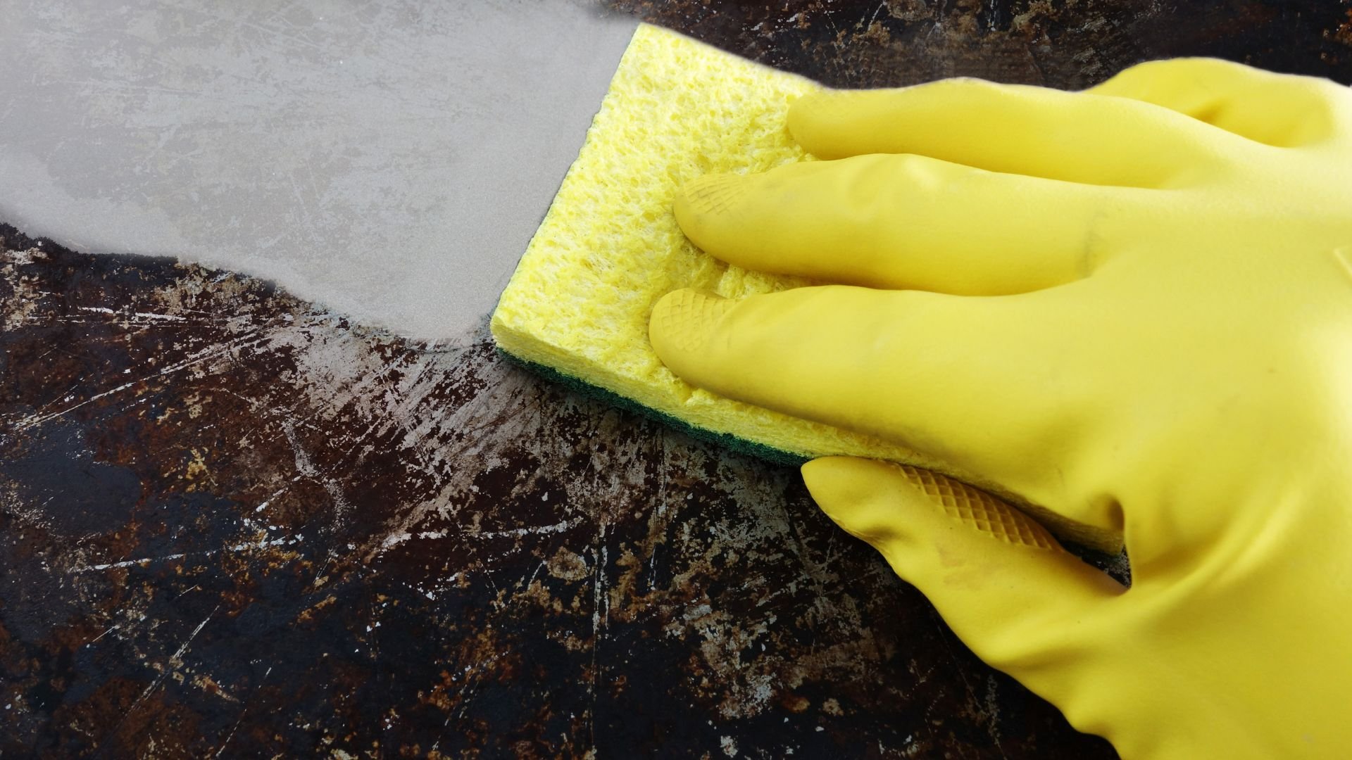 Yellow gloved hand cleaning dirty surface with yellow sponge
